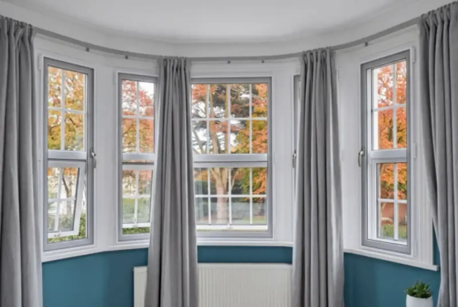 Bay window with slim white secondary glazing frames, grey curtains and autumn trees beyond