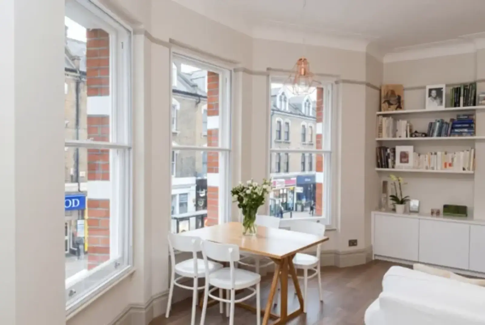 Period living room with three sash windows, a dining table and bookshelves overlooking a high street